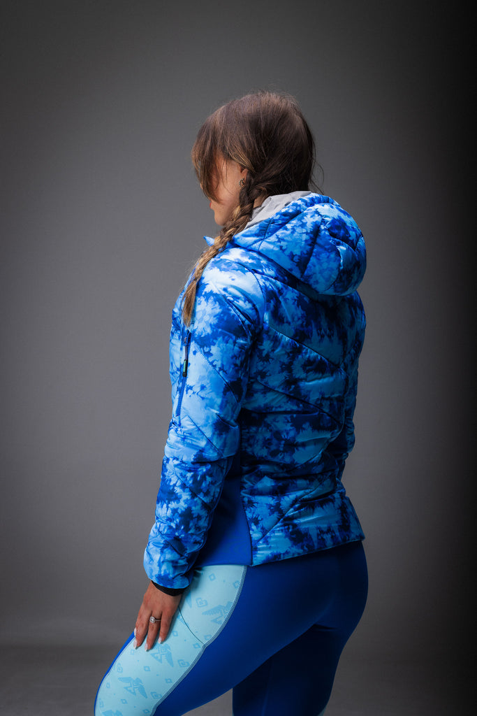 A person wearing a blue tie-dye patterned puffer jacket with zipped pockets, standing in a natural outdoor setting.