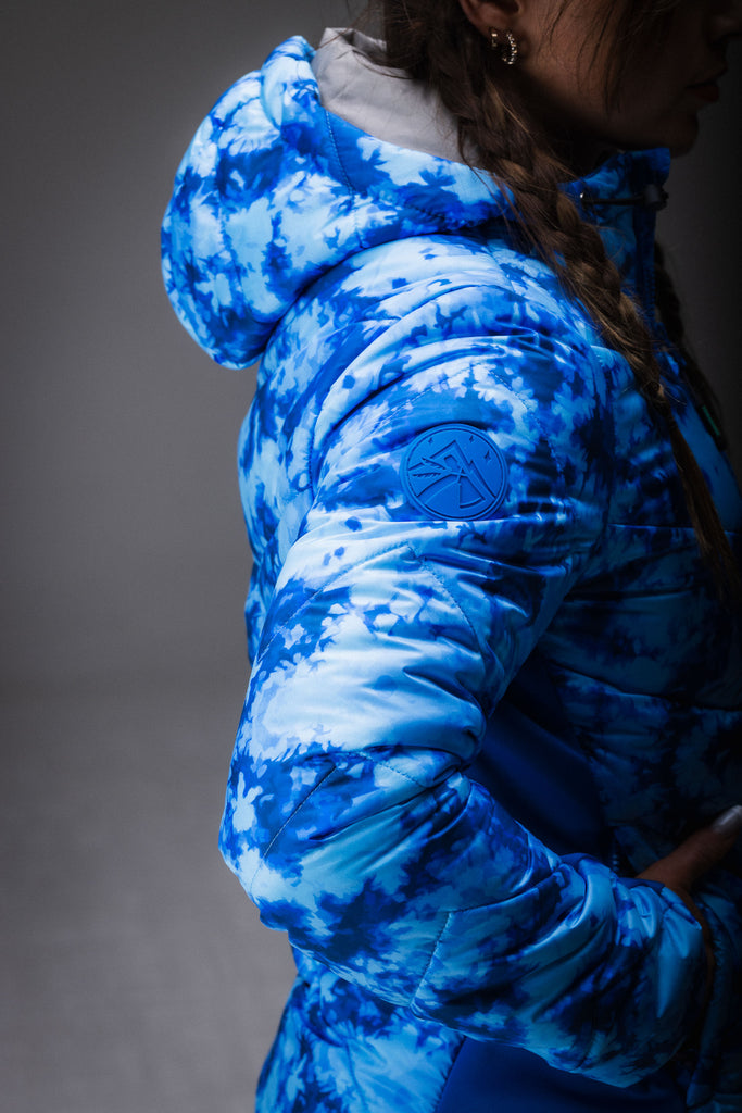 A person wearing a blue tie-dye patterned puffer jacket with zipped pockets, standing in a natural outdoor setting.