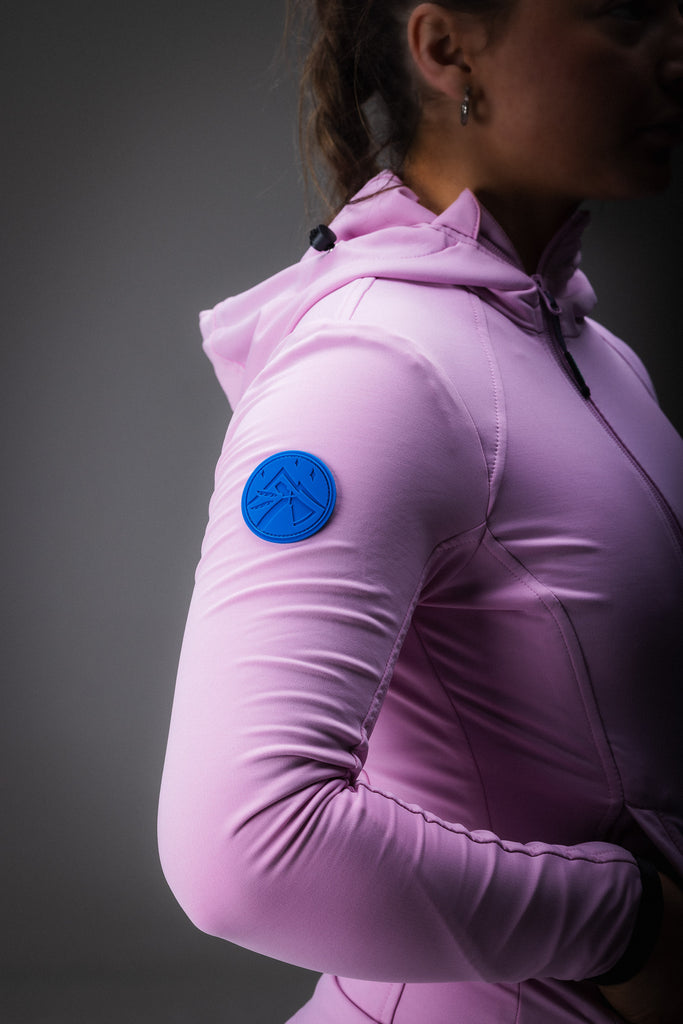 A person wearing a pink midlayer with a hood, zipped pockets, and an off-centred zip fastener, paired with patterned leggings and socks.