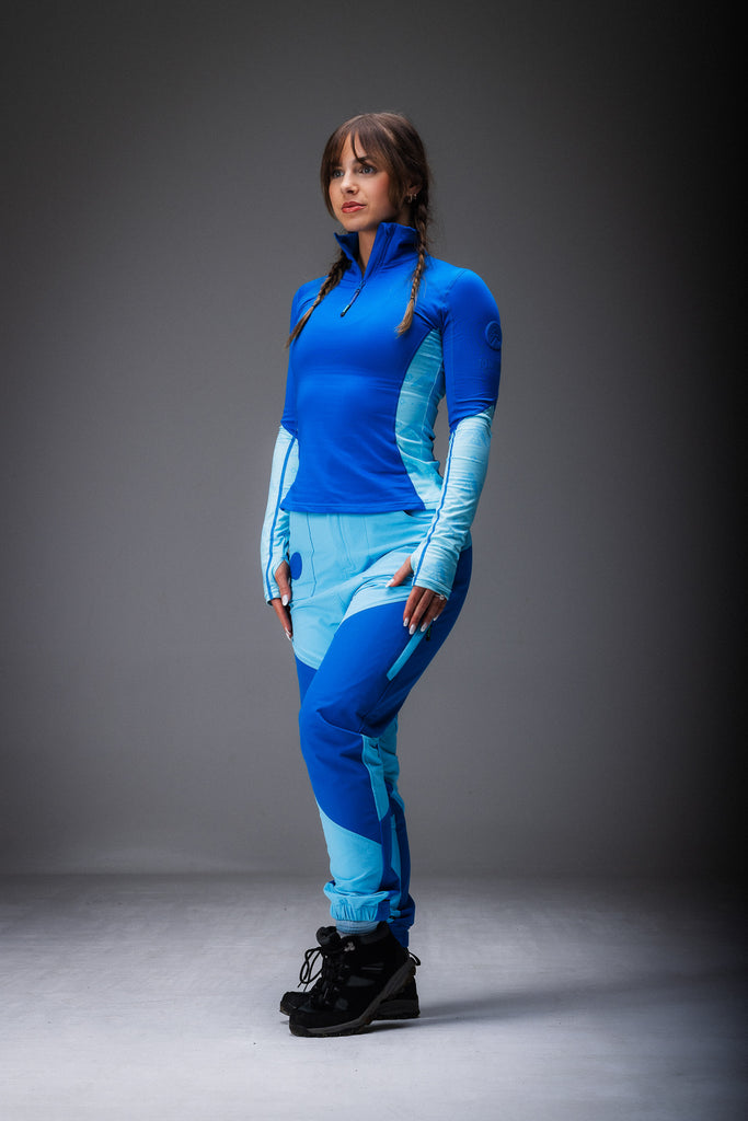 A woman standing, wearing high-waisted blue hiking pants with reinforced waterproof panels at the knee and seat, multiple pockets, and DWR coating.