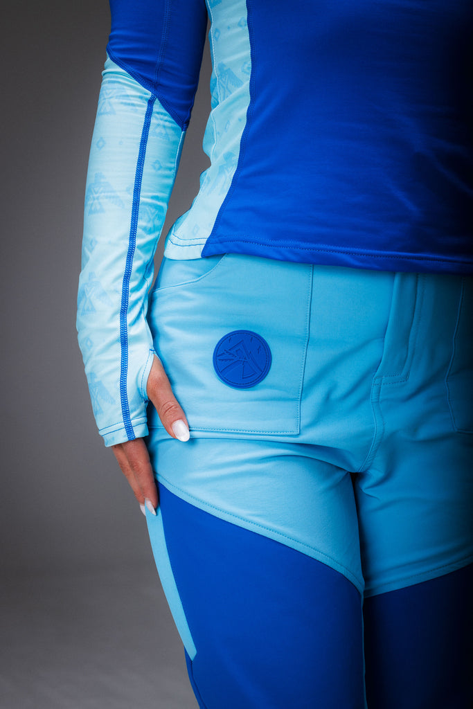 A woman standing, wearing high-waisted blue hiking pants with reinforced waterproof panels at the knee and seat, multiple pockets, and DWR coating.