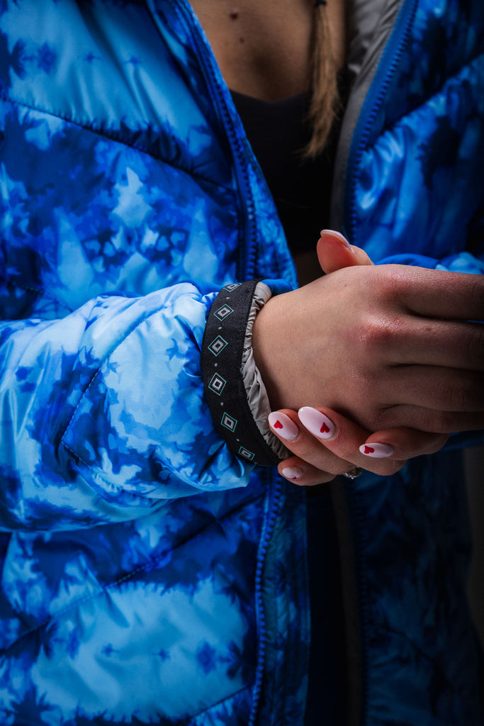 A person wearing a blue tie-dye patterned puffer jacket with zipped pockets, standing in a natural outdoor setting.
