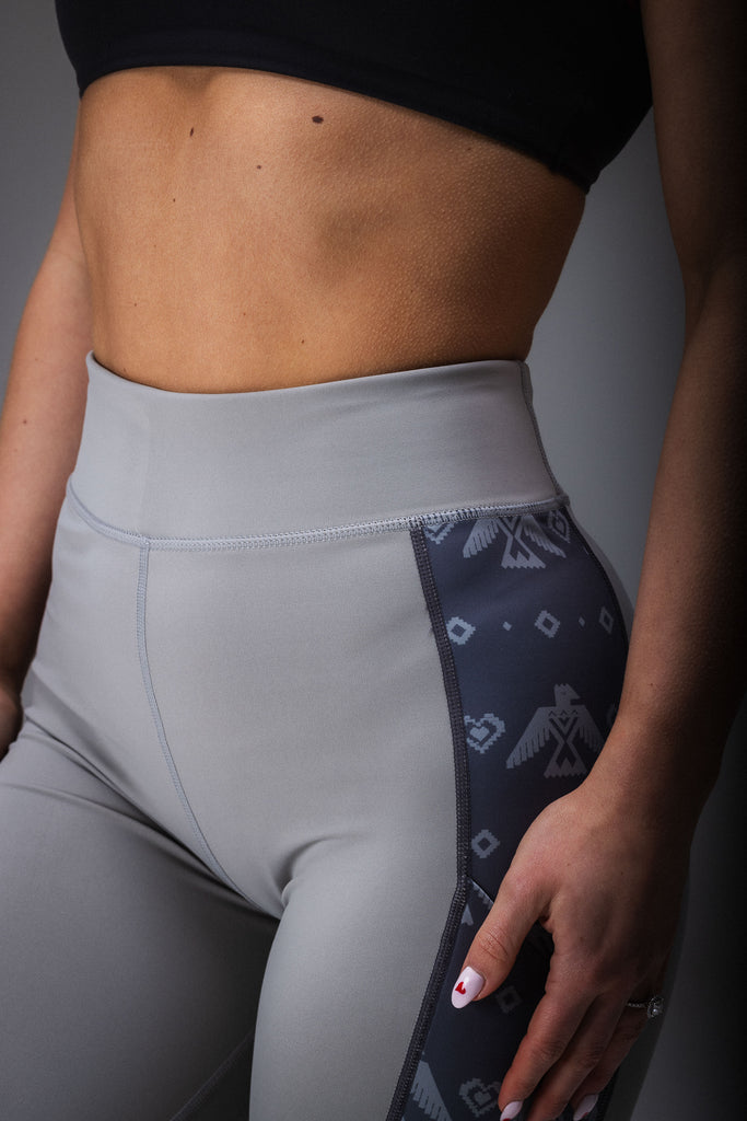 A person standing in a studio setting, wearing gray leggings with a geometric pattern, including black accents that resemble a tribal design.