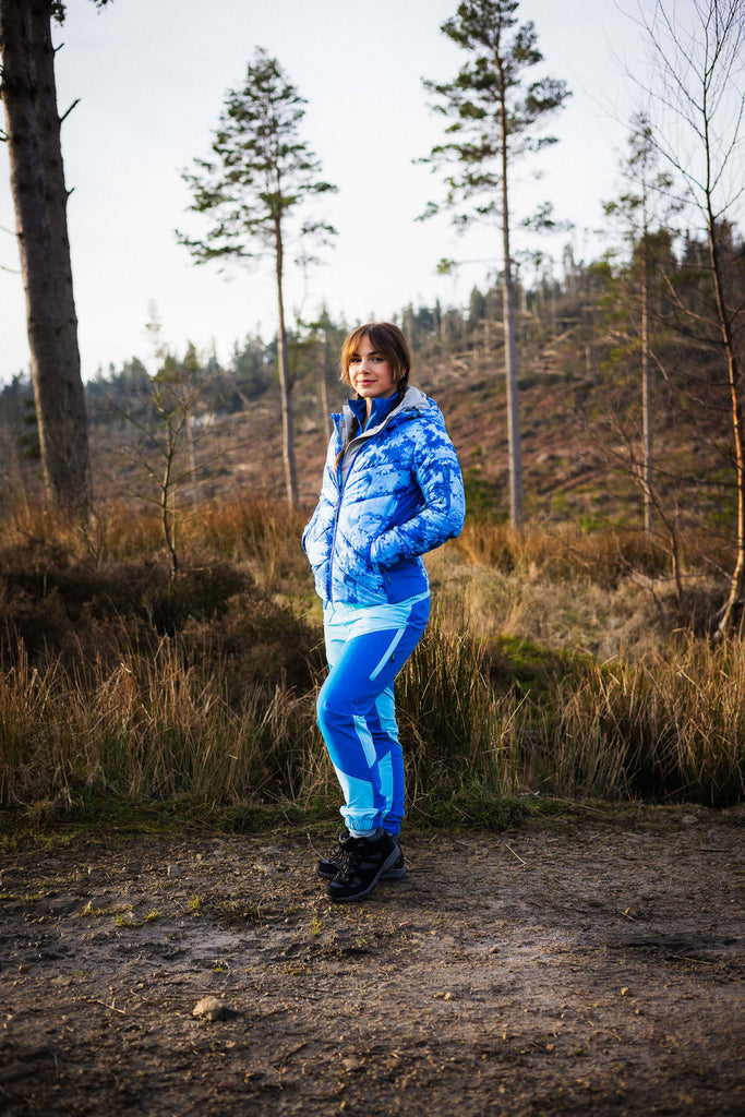 A person wearing a blue tie-dye patterned puffer jacket with zipped pockets, standing in a natural outdoor setting.