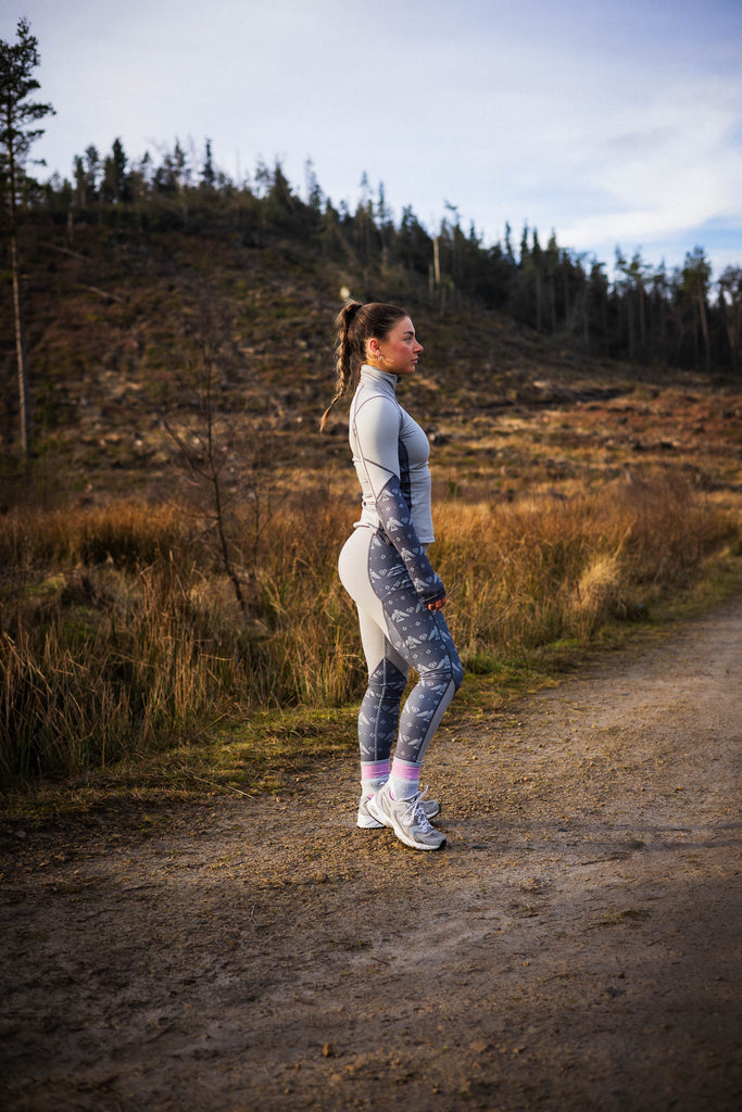 A person standing in a studio setting, wearing gray leggings with a geometric pattern, including black accents that resemble a tribal design.