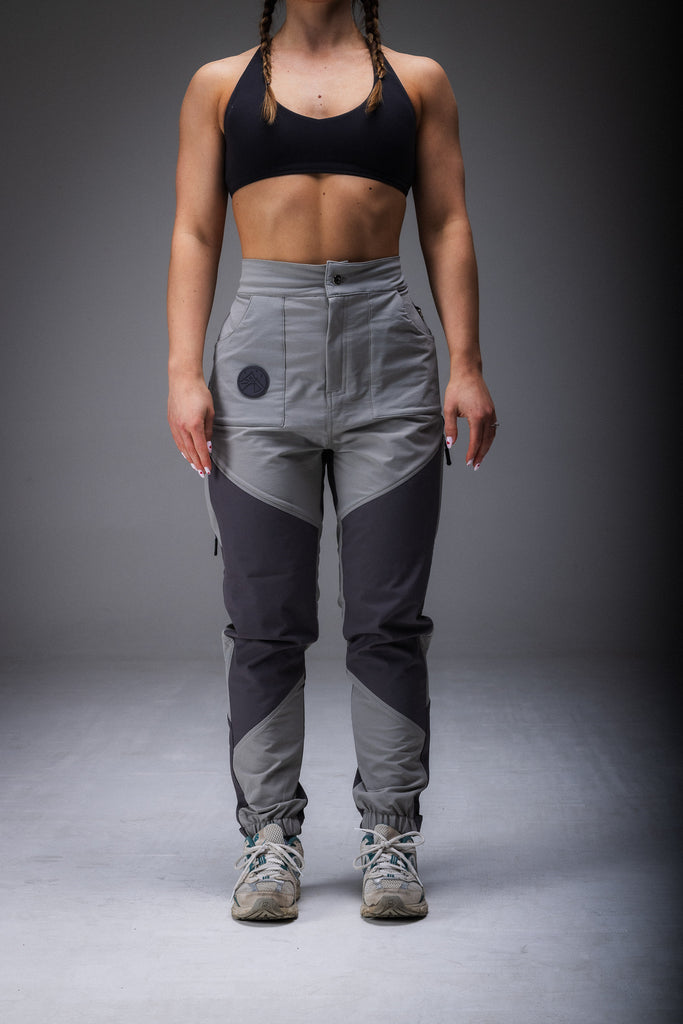 A woman standing, wearing high-waisted grey pants with reinforced waterproof panels at the knee and seat, suitable for hiking.