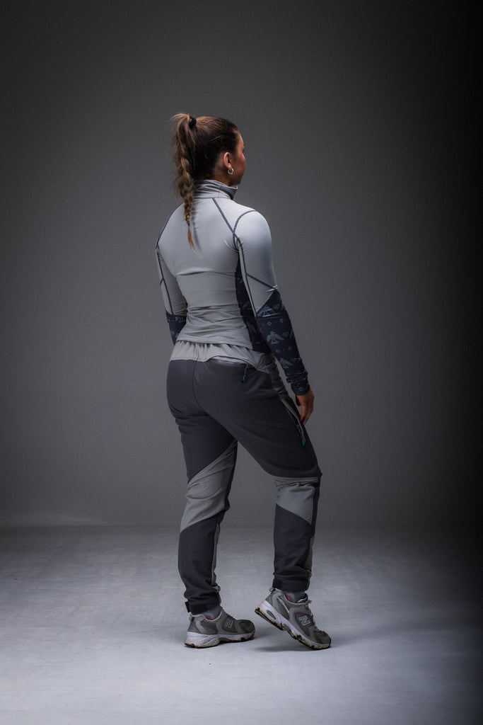 A woman standing, wearing high-waisted grey pants with reinforced waterproof panels at the knee and seat, suitable for hiking.