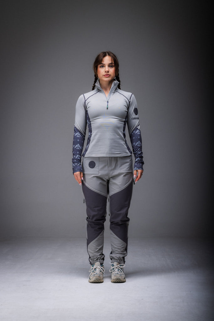 A woman standing, wearing high-waisted grey pants with reinforced waterproof panels at the knee and seat, suitable for hiking.