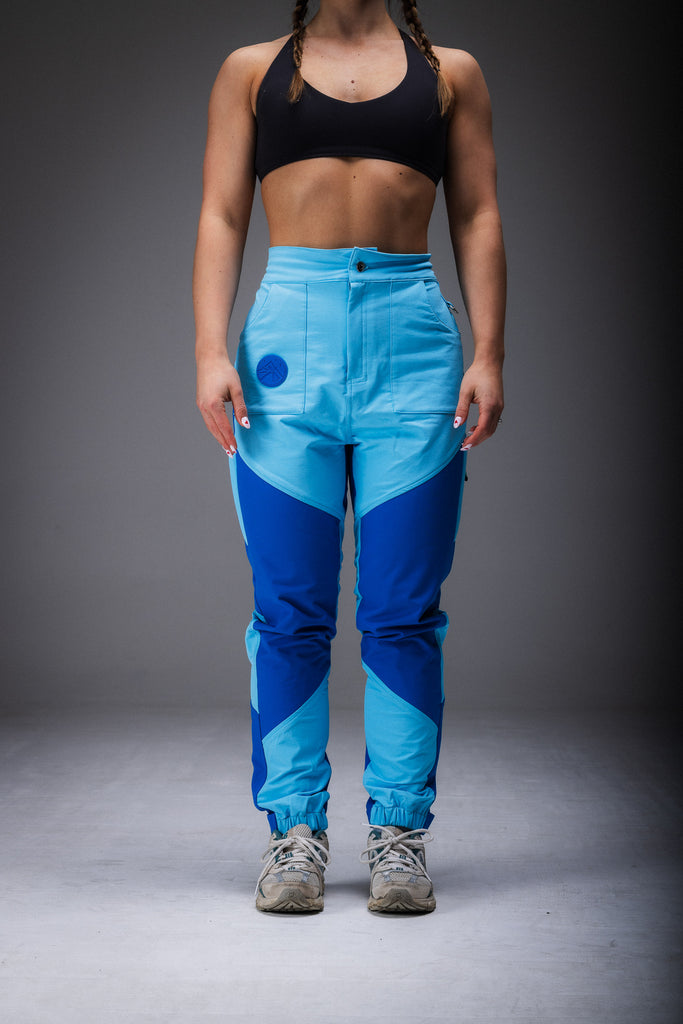 A woman standing, wearing high-waisted blue pants with reinforced waterproof panels at the knee and seat, multiple pockets, and Velcro around the ankles.