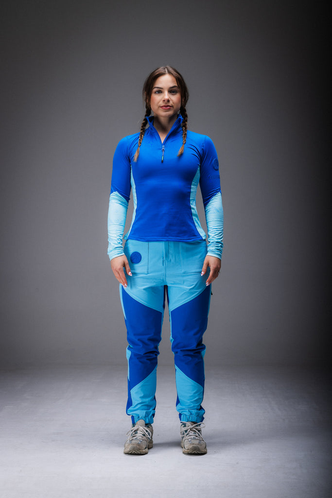A woman standing, wearing high-waisted blue pants with reinforced waterproof panels at the knee and seat, multiple pockets, and Velcro around the ankles.